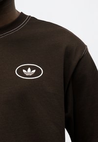 Σκούρο καφέ φούτερ με λευκό οβάλ λογότυπο που παρουσιάζει σχέδιο τριφυλλιού στο πάνω μέρος του στήθους, ορατή λευκή ραφή στο γιακά και στον ώμο.