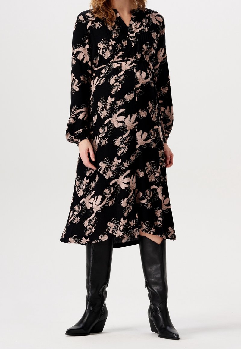 Femme portant une robe midi noire à imprimé floral rose et manches longues, associée à des bottes noires montantes à talons.