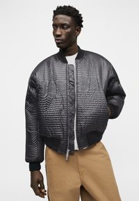 Άνδρας που φοράει μαύρο μπουφάν bomber με μεγάλα ανάγλυφα γράμματα, λευκό πουκάμισο και καφέ παντελόνι, στέκεται μπροστά σε ένα λευκό φόντο.