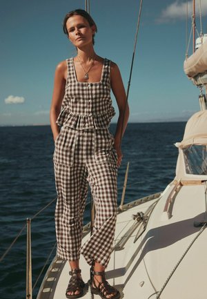 Kvinde stående på dæk af en sejlbåd iført ærmeløs ternet jumpsuit og brune sandaler, med hav og klar himmel i baggrunden.