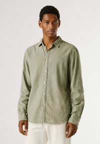 Νέος άνδρας όρθιος, φοράει ανοιχτοπράσινο πουκάμισο με κουμπιά και μακριά μανίκια και λευκό παντελόνι, με ουδέτερο φόντο.