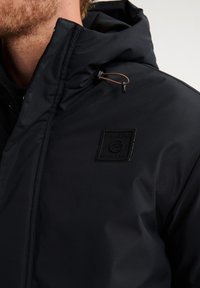 Giacca nera con cappuccio realizzata in tessuto liscio. Presenta una toppa con logo davanti, cordini regolabili sul cappuccio e un design con colletto alto.
