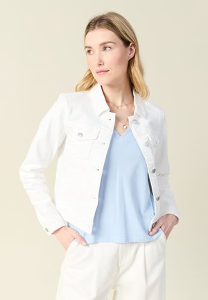 Weiße Jeansjacke mit silberner Hardware, zwei Fronttaschen und kragenfreiem Kragen. Getragen über einem hellblauen V-Ausschnitt-Oberteil und weißen Hosen.