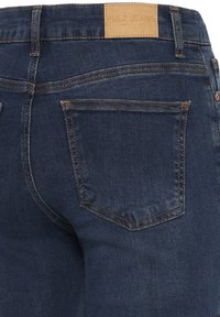 Vue arrière d'un jean en denim bleu foncé avec des coutures marron, une poche sur la droite et une étiquette de marque en cuir marron clair sur la ceinture.