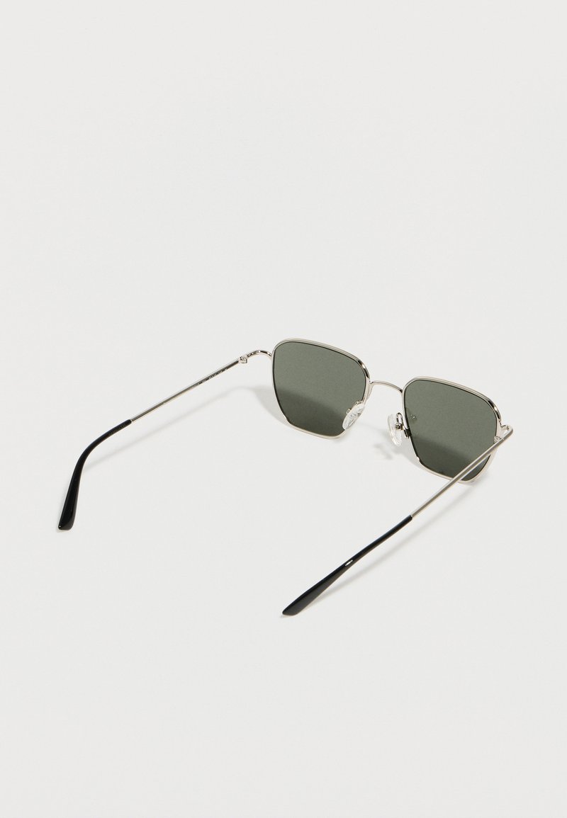 Lunettes de soleil à monture argentée avec des verres rectangulaires foncés et des embouts en plastique noir sur des branches métalliques fines, reposant sur une surface blanche.