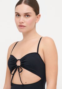 Maillot de bain noir avec de fines bretelles, devant froncé et côtés découpés. Il présente un détail de bague central et des liens en bas, texture lisse.