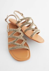 Sandali slingback con cinturini in tessuto dorato testurizzato, suole piatte in tono marrone e design a punta chiusa. I cinturini presentano un motivo intrecciato per un tocco di dettaglio in più.