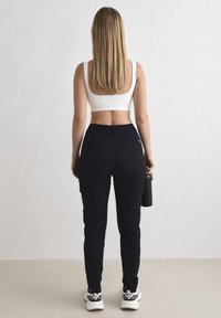 Femme aux cheveux blonds longs, de dos face à un mur, portant un soutien-gorge de sport blanc, un pantalon noir, des baskets blanches, tenant une bouteille d'eau noire dans la main droite.