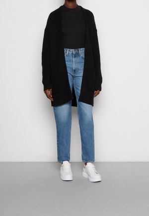 Personne portant un long cardigan noir, un haut noir, un jean taille haute bleu et des baskets blanches, debout devant un fond uni.
