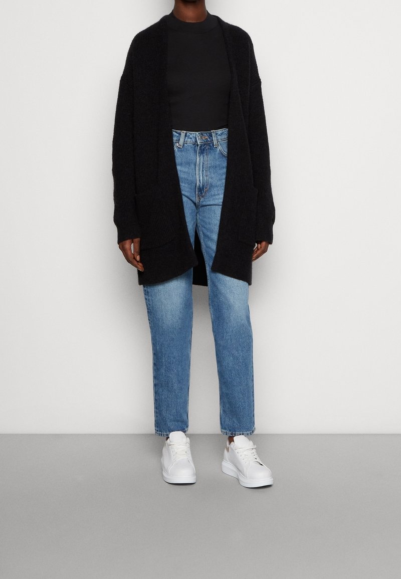 Personne portant un long cardigan noir, un haut noir, un jean taille haute bleu et des baskets blanches, debout devant un fond uni.