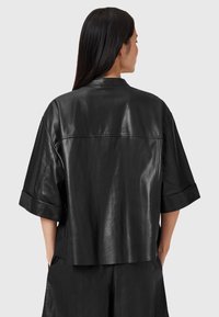 Black leather short-sleeve shirt with a relaxed fit, featuring a small collar, wide sleeves, and visible stitching details at the back and seams.