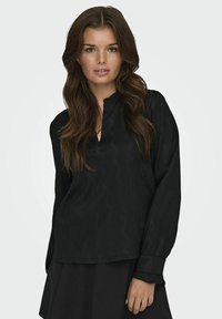 Schwarze Bluse mit langen Ärmeln aus strukturiertem Stoff, V-Ausschnitt und taillierten Manschetten. Kombiniert mit einem schwarzen Rock, was einen schichtenhaften Look erzeugt.