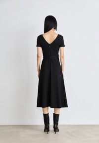 Kobieta z prostymi czarnymi włosami stoi twarzą do ściany, ubrana w czarną sukienkę midi z dekoltem w kształcie V i czarne bootsy na obcasie, na betonowej podłodze.