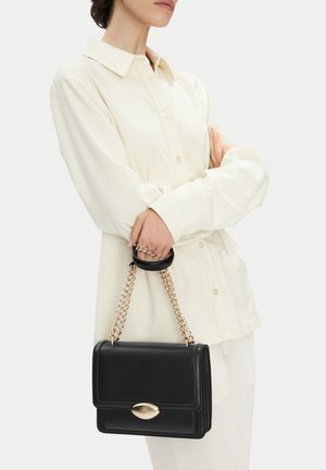 Donna con camicia cremisi a maniche lunghe abbottonata, che tiene una borsa in pelle nera con tracolla a catena dorata e chiusura ovale dorata all'altezza della vita.