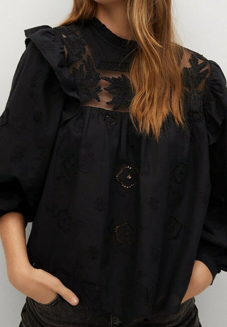 Blouse noire avec empiècement brodé transparent, épaules à volants, manches bouffantes et motifs floraux portée par une personne aux longs cheveux châtain clair.