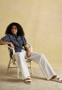 Tumedaga mustritega lühikeste varrukatega särk, valged lina püksid ja heledad sandaalid. Istumas rattanist toolil tekstuurse beež tumba taustal.