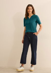 Top corto de color verde azulado con capucha, que presenta un corte holgado. Combinado con unos jeans de color azul oscuro, ligeramente acampanados, y zapatillas blancas con suela amarilla.