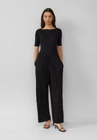 Haut noir à manches courtes associé à un pantalon noir taille haute plissé. Le pantalon présente des poches latérales. Le modèle porte des talons ouverts noirs.