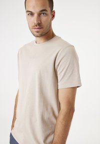 Camiseta de algodón beige con mangas cortas, cuello redondo, bordado sutil del logo en el delantero y textura suave. Diseño de corte clásico.