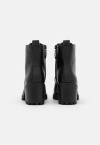 Bottes courtes noires en matière synthétique lisse, avec un talon épais, une fermeture éclair et un détail de marque en relief sur le talon.