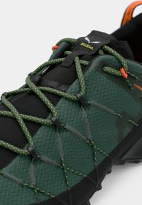 Sapato de caminhada verde com parte superior em malha texturizada, detalhes em preto e atacadores verdes refletivos. Inclui sola de borracha preta e um logótipo proeminente.