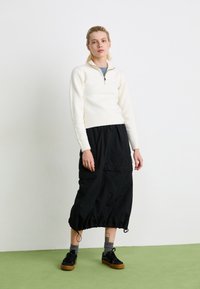 White knit sweater with a half-zip collar, paired with a black elasticated skirt. Black sneakers and grey socks complete the outfit.