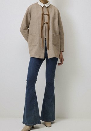 Personne portant une veste beige avec des liens à l'avant, une chemise blanche à col, un jean évasé bleu foncé et des chaussures à talons beiges, debout devant un fond uni.