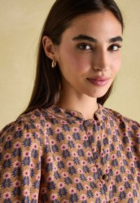 Braun gemusterte Bluse mit floralen Motiven in Pink und Grün. Rundhalsausschnitt und Knopfdetails. Glattes, weiches Material mit lockerer Passform.