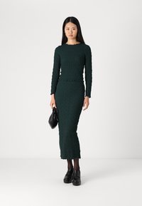 Green, textured midi dress with long sleeves and scalloped neckline; paired with black loafers and a small bag.
