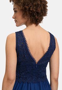 Tengerész-kék ruha, virágmintás csipkebetéttel, mély V-hátkivágással, fodros szélekkel és lágyan omló szoknyával, hangsúlyozva a textúrát és a formatervezést.