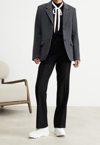 Grå ullblazer med reva laplar, i kombination med svarta byxor och en svart blus med en vit slips. Vita sneakers fulländar looken.