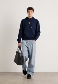Felpa navy con tasca frontale e cordini, abbinata a jeans larghi di un blu chiaro. Il modello porta una borsa nera e indossa scarpe nere.