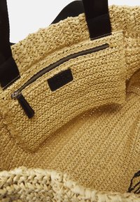 Intérieur d'un sac fourre-tout tissé naturel avec poignées en tissu noir et poche zippée dotée d'une tirette en cuir noir et d'une étiquette logo.