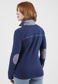 Chemise à manches longues bleu marine avec un col à motifs, des coudières fleuries et des poignets boutonnés ; présente un design ajusté et des coutures décoratives.