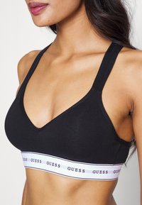 Svart bralette med dypt V-halsdesign, laget av mykt stoff, med brede stropper og et hvitt bånd merket med "GUESS."