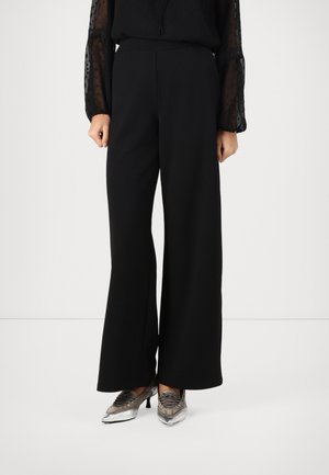Frau trägt schwarze Hosen mit weitem Bein, transparentes schwarzes Langarmtop und silberne spitze High-Heel-Schuhe und steht auf weißem Boden.