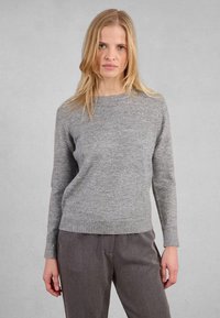 Donna con capelli biondi che indossa un maglione grigio a maniche lunghe e pantaloni larghi grigio scuro, in piedi contro uno sfondo grigio chiaro uniforme.