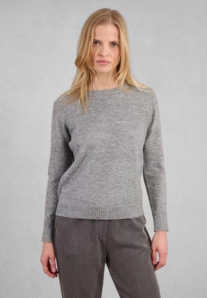 Donna con capelli biondi che indossa un maglione grigio a maniche lunghe e pantaloni larghi grigio scuro, in piedi contro uno sfondo grigio chiaro uniforme.