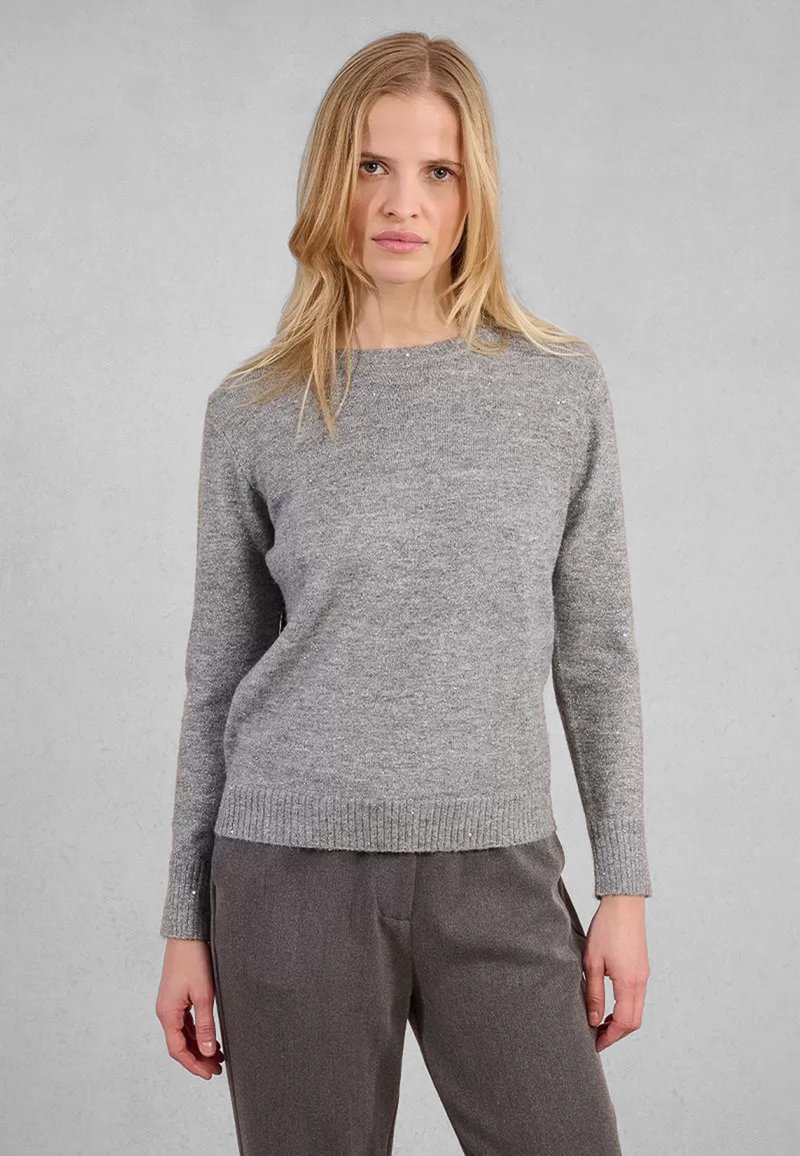 Donna con capelli biondi che indossa un maglione grigio a maniche lunghe e pantaloni larghi grigio scuro, in piedi contro uno sfondo grigio chiaro uniforme.