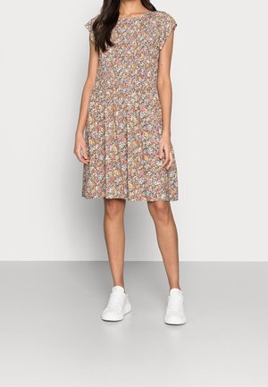 Kobieta w sukience w kwiaty bez rękawów sięgającej kolan i białych trampkach, stojąca na tle jednolitego jasnoszarego tła.