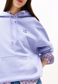 Lila hoodie met een kangaroozak, voorzien van een klein blauw-wit logo. Draagt over een blauw-roze geruit overhemd, wat een gelaagde uitstraling geeft.