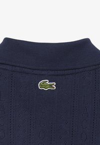 Mørkeblå strikket polo med krave og subtilt mønster, prydet med et grønt broderet krokodillelogo. Glat tekstur og ribbede kanter.