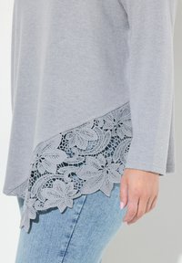 Grå langærmet top med blonder i kanten, der har en tekstureret overflade. Kombineret med lyseblå denim jeans.