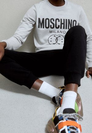 Person, der sidder på gulvet iført en hvid Moschino Milano sweatshirt, sorte bukser, hvide sokker og farverige sneakers med orange, grønne og metalliske detaljer.