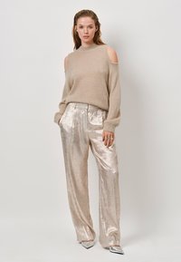 Beige gebreide trui met een off-shoulder ontwerp en lange mouwen, gecombineerd met glanzende zilveren wijde broek en metallic zilveren schoenen.