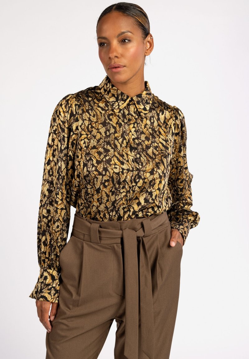 Bruine blouse met een patroon en gouden accenten, voorzien van een kraag en lange mouwen. Gecombineerd met bruin, hoge taille broek met brede pijpen.