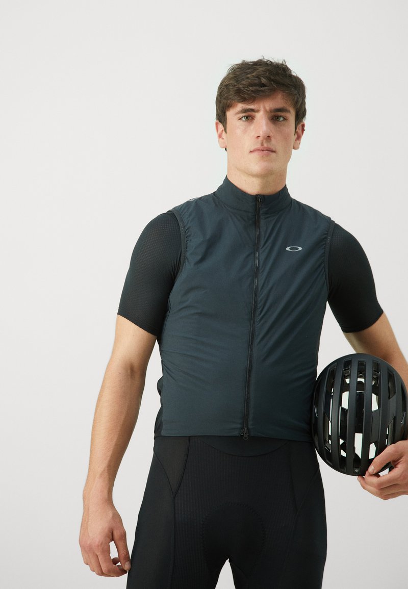 Schwarze Radsportweste mit hohem Kragen und durchgehendem Reißverschluss, kombiniert mit einem schwarzen kurzärmeligen Baselayer und schwarzen Radshorts. Helme in der Hand.