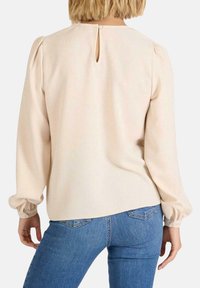Blusa de manga larga en suave tejido beige, con detalle de corte en la parte posterior y mangas abullonadas, combinada con jeans ajustados azules.