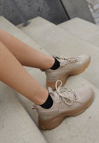 Beige sportsko med en tekstureret overdel, glitrende accenter og en tyk, gennemsigtig sål, parret med sorte glitrende sokker.