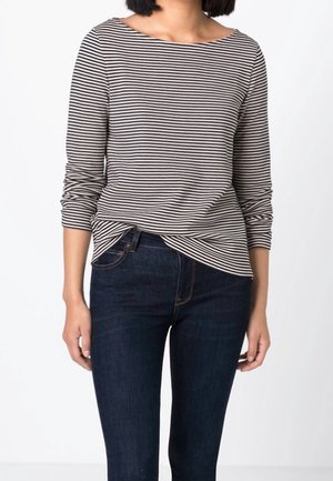 Mujer con una camiseta de manga larga a rayas blancas y negras metida en unos vaqueros azul oscuro, de pie frente a un fondo blanco.